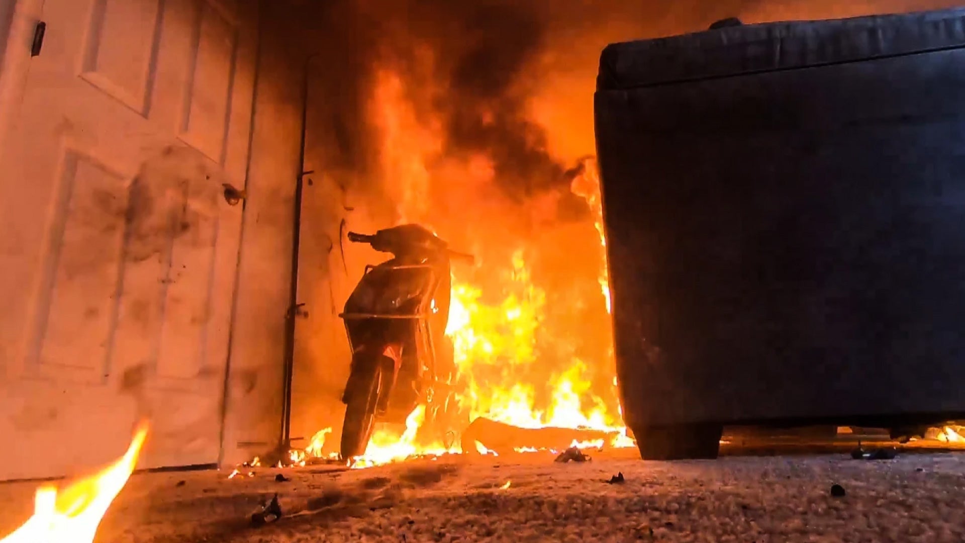 Scooter engulfed in flames, indoor charging, with a large couch nearby.