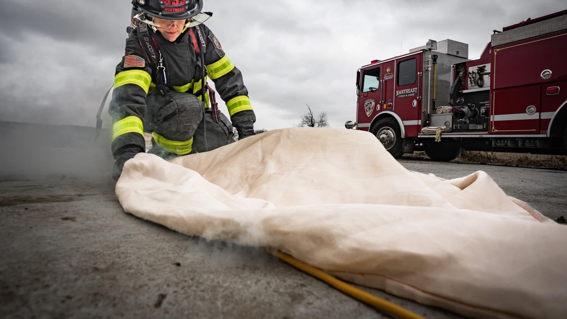 Firefighter using a fire blanket on a smoldering object with a fire truck in the background.