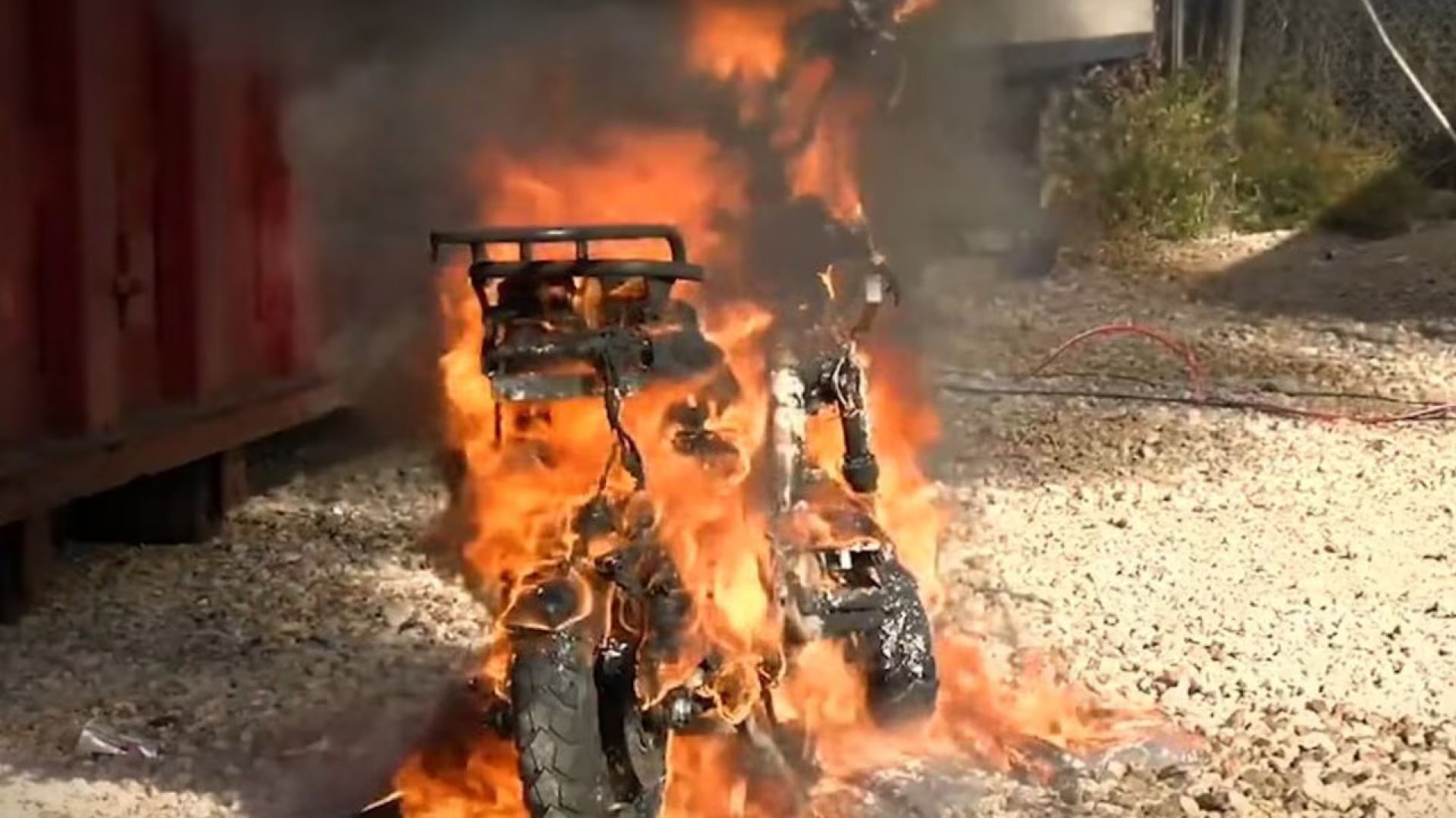 Motorcycle engulfed in flames on a gravel surface with a red container in the background.