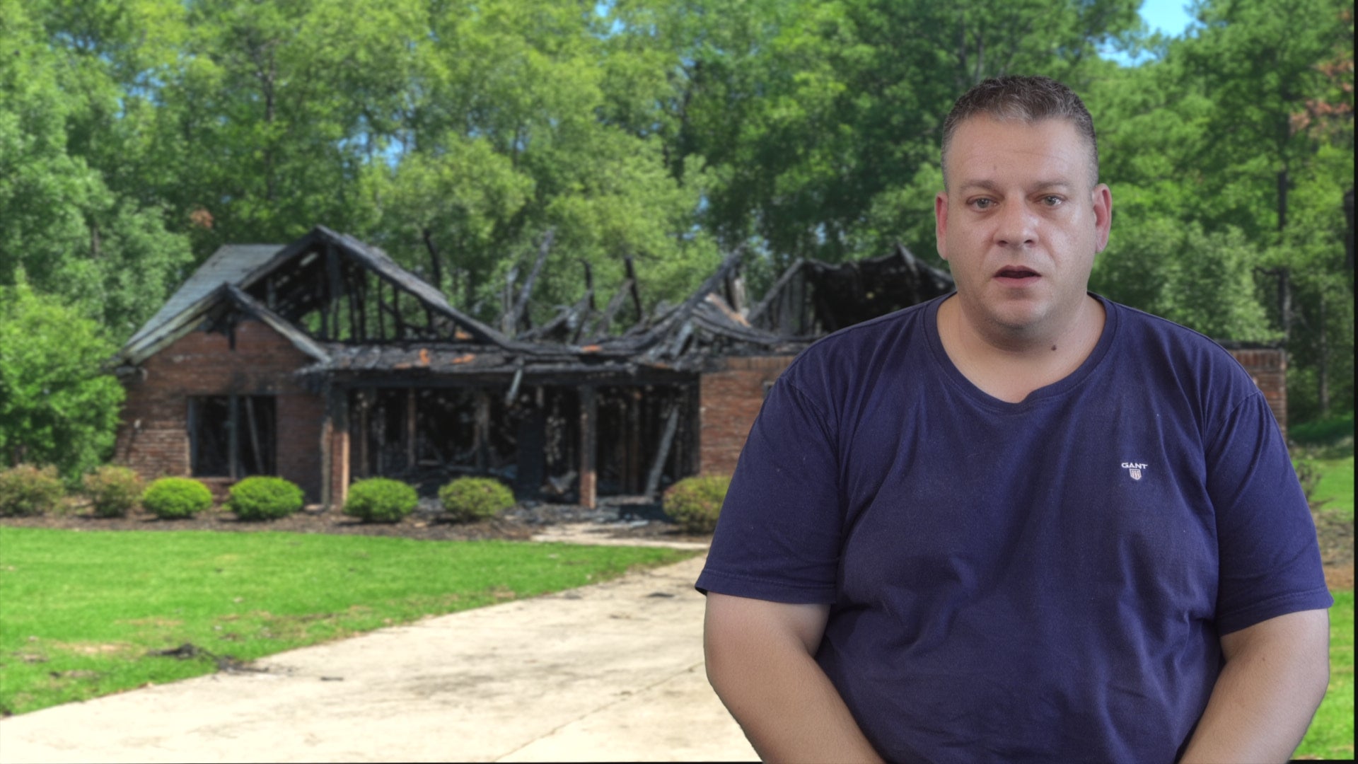 Load video: A video still of a man standing in front of the charred, blackened remains of a brick house destroyed by fire, appearing solemn and overwhelmed as he surveys the damage.