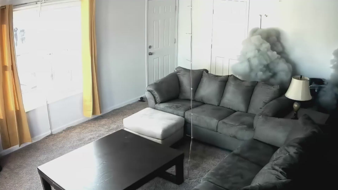A security camera video still of a living room where thick grey smoke is billowing from a device on a side table next to a sofa, illustrating the rapid onset of a lithium battery fire event.